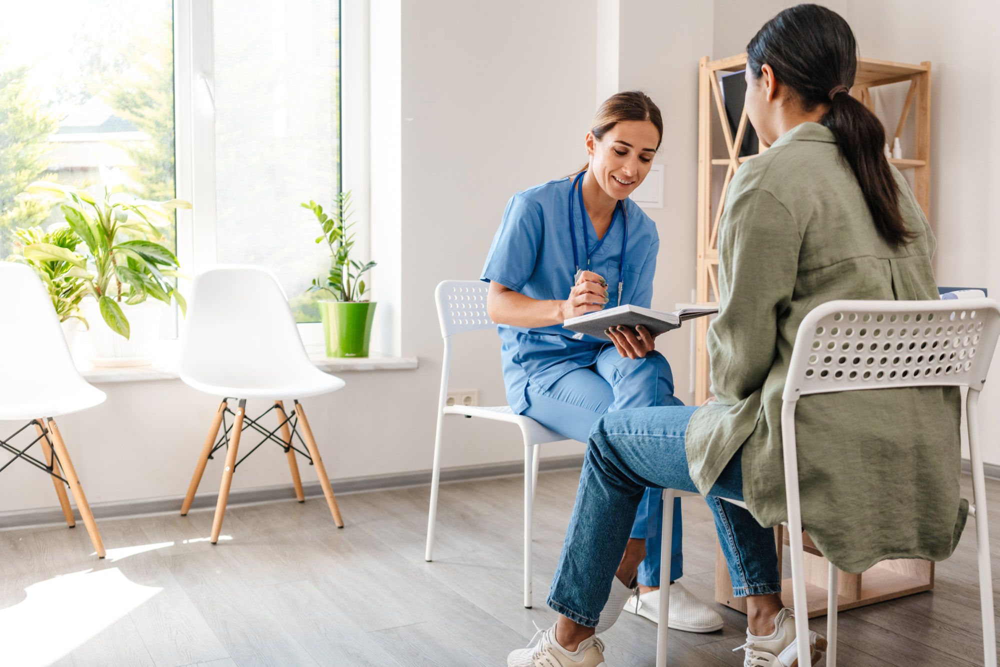 A nurse in blue scrubs sitting with a patient in a medical office, taking notes in a notebook. The room is bright and modern, with potted plants, white chairs, and large windows letting in natural light.